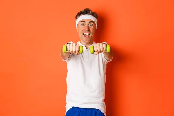 Képek aktív középkorú fitness srác, csinál sport gyakorlatok súlyzók és mosolygós izgatott, áll a narancs háttér felett — Stock Fotó