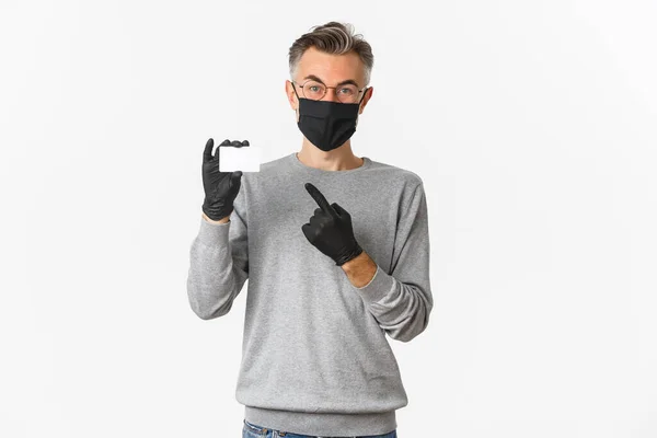 Концепция ковид-19, социальное дистанцирование и образ жизни. Красивый мужчина средних лет в медицинской маске, перчатках и очках, указывающий пальцем на кредитку, стоящий на белом фоне — стоковое фото