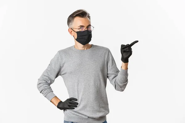 Концепция коронавируса, образа жизни и карантина. Изображение мужчины средних лет в очках, медицинская маска и перчатки, указывающие пальцем вправо, показывающие промо-баннер, стоящий на белом фоне — стоковое фото