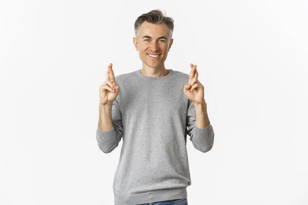 Imagem do esperançoso homem de meia-idade fazendo um desejo, cruzando os dedos e sorrindo, antecipando boas notícias, de pé sobre fundo branco — Fotografia de Stock