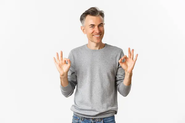 Retrato de homem de meia-idade bonito, sorrindo e olhando confiante ao mostrar sinais ok, garantir que algo é bom, de pé sobre fundo branco — Fotografia de Stock