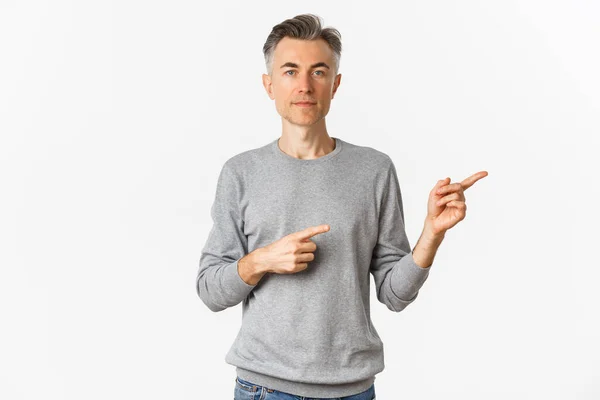 Πορτρέτο του ελκυστικού μεσήλικα άνδρα σε γκρι πουλόβερ, δείχνοντας τα δάχτυλα δεξιά και φαίνεται σοβαρή, δείχνει μια διαφήμιση, στέκεται πάνω από το λευκό φόντο — Φωτογραφία Αρχείου