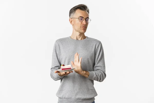 Retrato de homem de meia-idade descontente, olhando para longe nojento e rejeitando pedaço de bolo, de pé incomodado sobre fundo branco — Fotografia de Stock