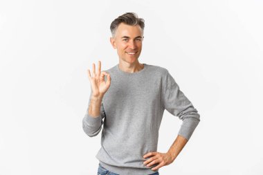 Gri süveterli yakışıklı orta yaşlı bir adamın resmi, tamam işareti gösteriyor ve gülümsüyor tatmin olmuş, ürün öneriyor, onaylıyor ve onaylıyor, beyaz arka planda duruyor.