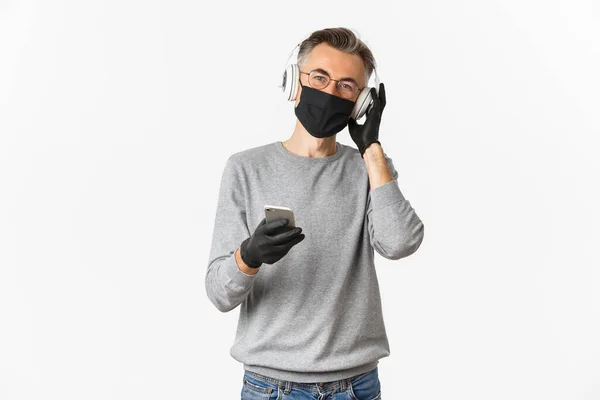 Concept of covid-19, társadalmi távolság és életmód. Kép középkorú férfi orvosi maszk, kesztyű és szemüveg, zenét hallgatni a fejhallgató és gazdaság okostelefon — Stock Fotó