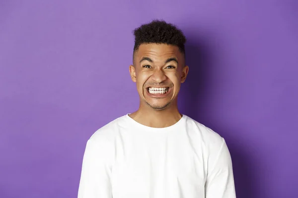 Primer plano del gracioso afroamericano con sudadera blanca, apretando los dientes y haciendo muecas ante la cámara, sacando una sonrisa forzada, de pie sobre el fondo púrpura —  Fotos de Stock