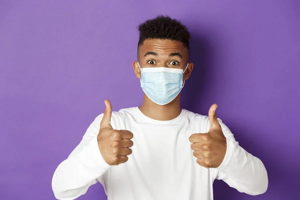 Concept of coronavirus, quarantine and social distancing. Close-up of young african-american guy in medical mask, looking excited and recommending something, showing thumbs-up in approval — Stock Photo, Image