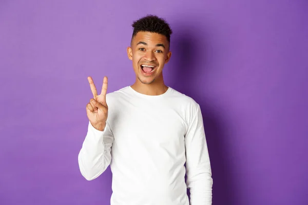 Bild von fröhlichen afrikanisch-amerikanischen Kerl in weißem Sweatshirt, zeigt zwei Finger und lächelt, steht über lila Hintergrund — Stockfoto