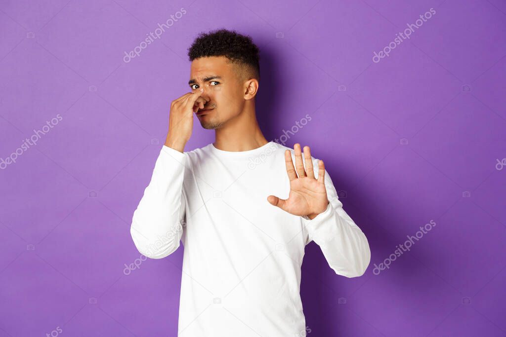 Image of african-american guy rejecting something with awful smell, shut nose from stink and showing stop gesture, decline offer, standing over purple background