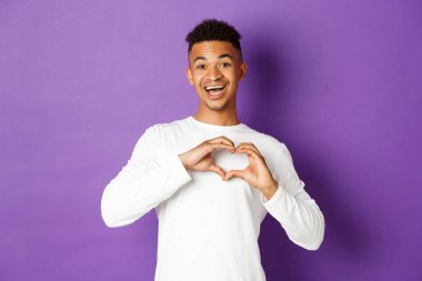 Sevimli Afro-Amerikalı erkek arkadaşın görüntüsü, gülümseyen ve kalp işareti gösteren, aşkını itiraf eden, mor arka planda duran