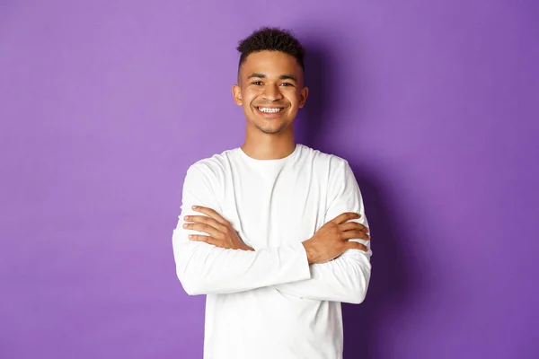 Portrait of handsome african-american male student in white sweatshirt, cross arms on chest and smiling confident, standing pleased over purple background — Stock Photo, Image