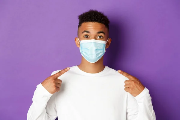 Concept of coronavirus, quarantine and social distancing. Close-up of young african-american man smiling, recommending to wear medical mask during covid-19, standing over purple background — Stock Photo, Image