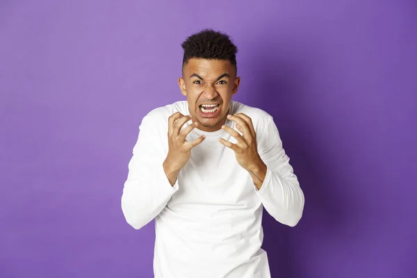 Image de jeune homme afro-américain en colère, criant et serrant les poings de la haine, regardant fou et énervé à la caméra, debout sur fond violet — Photo