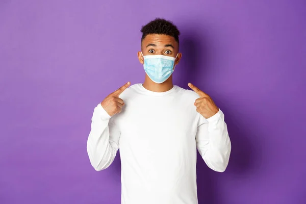 Concept de covie-19, pandémie et distanciation sociale. Image d'un homme afro-américain amusé, recommander de porter un masque médical pendant le coronavirus, debout sur fond violet — Photo