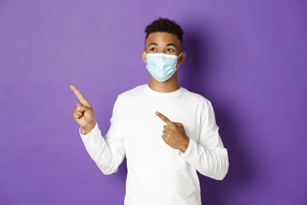 Concept de coronavirus, quarantaine et mode de vie. Beau jeune homme afro-américain en masque médical, pointant et regardant le coin supérieur gauche, montrant de la publicité, posant sur fond violet — Photo
