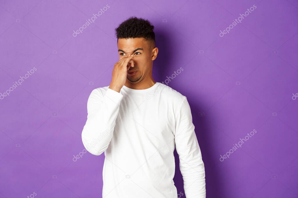 Portrait of young african-american guy in white sweatshirt, shut nose and grimacing from bad smell, looking at something nasty with disgusting reek, standing over purple background
