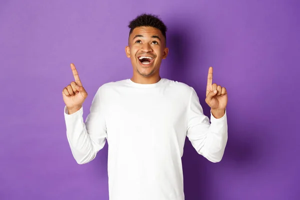 Schöner aufgeregter afrikanisch-amerikanischer Typ, in weißem Sweatshirt, erstaunt guckend und mit erhobenem Zeigefinger, supercoole Werbung zeigend, vor lila Hintergrund stehend — Stockfoto