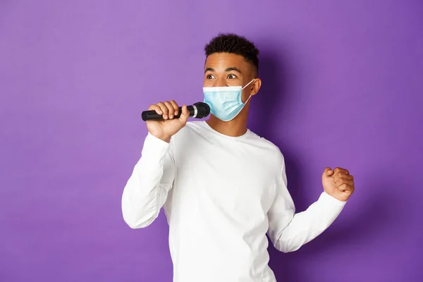 Έννοια του covid-19, πανδημία και κοινωνική αποστασιοποίηση. Χαρούμενος αφροαμερικάνος που διασκεδάζει στο καραόκε, φοράει ιατρική μάσκα και τραγουδάει στο μικρόφωνο, στέκεται πάνω από μωβ φόντο — Φωτογραφία Αρχείου