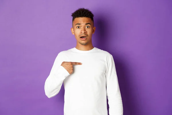 Portrait of confused and nervous african-american young man, pointing at himself, being chosen, standing over purple background — Stock Photo, Image