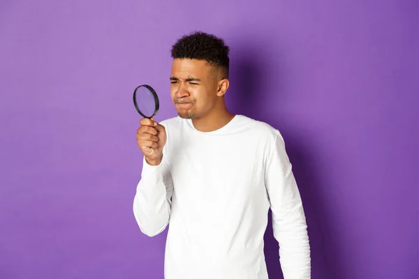 Immagine di giovane studente maschio afro-americano in cerca di qualcosa con lente d'ingrandimento, strizzando gli occhi come non si può vedere qualcosa di piccolo, in piedi su sfondo viola — Foto Stock