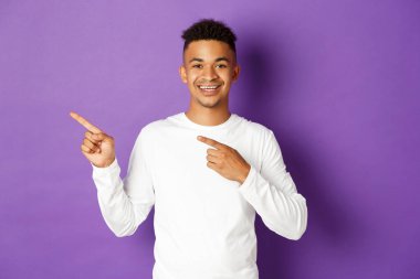 Beyaz kazaklı neşeli Afro-Amerikalı adam logo için fotokopi alanı gösteriyor, parmaklarını sola gösteriyor, kameraya gülümsüyor, mor arka planda duruyor.
