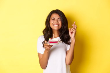 Şirin Afro-Amerikan kadın portresi, kapalı gözler ve gülümseme, doğum günü pastasında dilek tutmak için dua eden parmaklar, doğum gününü kutlayan, sarı arka planda duran