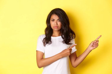 Şüpheci ve eğlendirilmemiş Afro-Amerikalı bir kızın portresi, kötü bir şeyi yargılamak, hayal kırıklığına uğramış bir ifadeyle parmaklarını işaret etmek, sarı arka planda durmak.