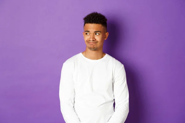 Image of disappointed and sad african-american man, looking left and grimacing, standing over purple background — Stock Photo, Image