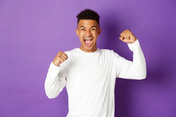 Beeld van een gelukkige Afrikaans-Amerikaanse man, wroeten voor het sportteam, handen omhoog steken en schreeuwen van vreugde, ja zeggen en de overwinning vieren, triomferen over paarse achtergrond — Stockfoto