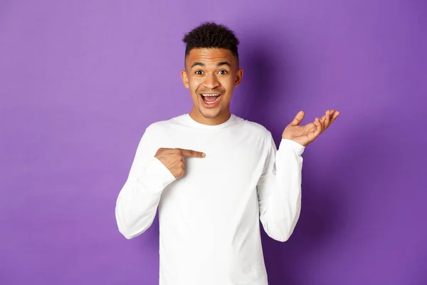 Imagen del hombre afroamericano sorprendido en sudadera blanca, apuntándose a sí mismo y levantando la mano sorprendido, sonriendo feliz, de pie sobre fondo púrpura —  Fotos de Stock