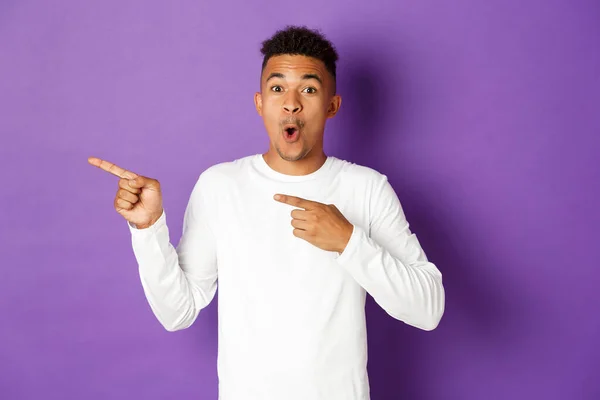 Porträt eines erstaunten afrikanisch-amerikanischen Mannes in weißem Sweatshirt, der beeindruckt aussieht und Werbung zeigt, mit dem Finger nach links zeigt, vor lila Hintergrund steht — Stockfoto