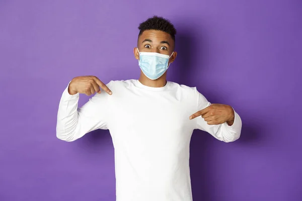Έννοια του coronavirus, καραντίνα και τρόπος ζωής. Ενθουσιασμένος αφροαμερικάνος με ιατρική μάσκα, να δείχνει το λογότυπο στο κέντρο και να δείχνει έκπληκτος, να στέκεται πάνω από μωβ φόντο — Φωτογραφία Αρχείου