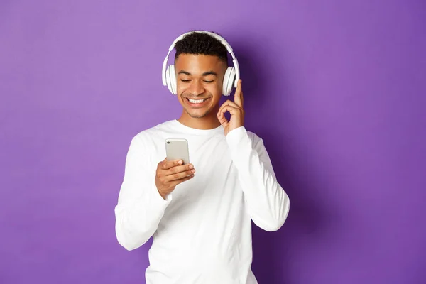 Beyaz kazaklı, kulaklıkla müzik dinleyen, cep telefonuna bakan, mor arka planda duran modern Afro-Amerikan bir adam. — Stok fotoğraf