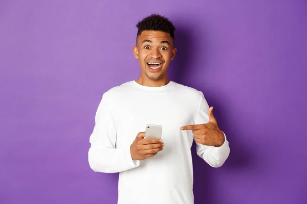 Imagen del hombre afroamericano asombrado, señalando con el dedo el teléfono móvil y sonriendo excitado, recomendando aplicación, de pie sobre el fondo púrpura —  Fotos de Stock