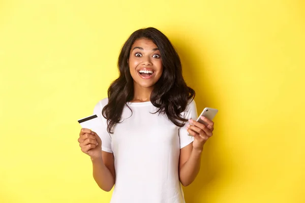 Porträtt av förvånad afrikansk-amerikansk flicka, håller smartphone och kreditkort, stående över gul bakgrund — Stockfoto
