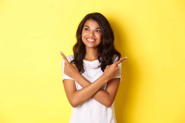 흰색 티셔츠를 입고 웃으며 왼쪽을 바라보고 있는 아프리카계 미국인 소녀의 매력적 인 모습, 두 개의 제안 사이에서 선택을 하는 동안 옆으로 손가락을 가리키며 노란 배경 위에 서 있는 모습 — 스톡 사진