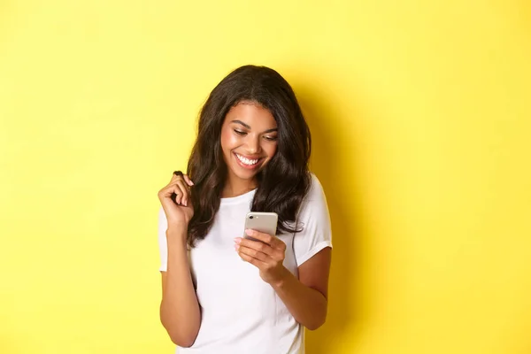 Imagen de la atractiva chica afroamericana en camiseta blanca, mensajería en el teléfono inteligente, mirando el teléfono móvil y sonriendo, de pie sobre el fondo amarillo — Foto de Stock