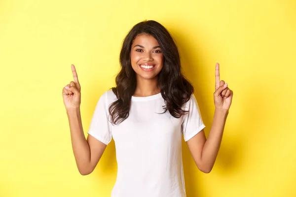 매력적 이고 자신감 있는 흑인 여성 모델의 사진 흰색 티셔츠를 입고 프모 로고를 손가락으로 가리키며 노란 배경 위에 서 있는 모습 — 스톡 사진