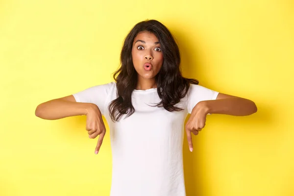 Retrato de menina afro-americana atraente, apontando os dedos para baixo e olhando espantado, mostrando algo curioso, de pé sobre fundo amarelo — Fotografia de Stock