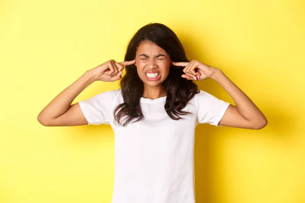 Portrait of annoyed and pissed-off african-american girl, shut her ears and grimacing from loud awful noise, being disturbed by something loud, standing over yellow background — 스톡 사진