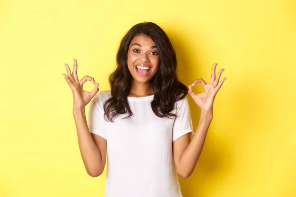 Portré boldog és elégedett afro-amerikai lány, fehér pólót visel, mutatja rendben jelek dicsérni kiváló választás, jóváhagyja és egyetért valamivel, áll a sárga háttér felett — Stock Fotó