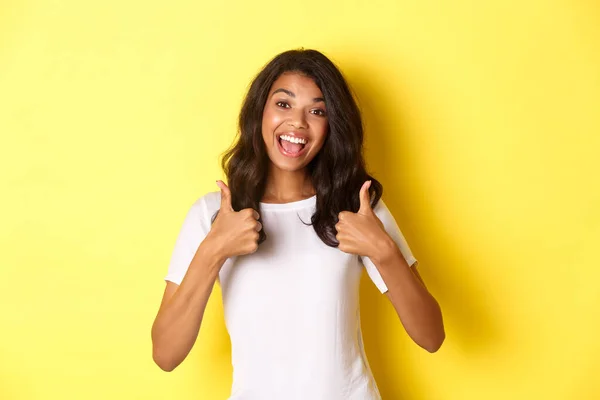 Retrato de menina afro-americana bonito e alegre, mostrando polegar-se em apoio, como e concordar com você, elogiando excelente trabalho, de pé sobre fundo amarelo — Fotografia de Stock