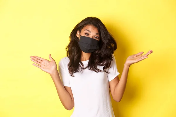 Il concetto di coronavirus, pandemia e stile di vita. Immagine di carina ragazza afro-americana in maschera, scrollando le spalle e cercando indiziato, non so nulla, in piedi su sfondo giallo — Foto Stock