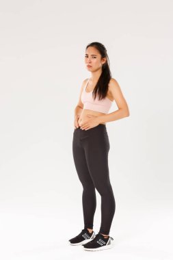 Kilo vermeye, karnına dokunmaya çalışan üzgün ve kasvetli Asyalı fitness kızı. Beyaz arka planda spor kıyafetleri içinde dururken idman ve diyet sonuçlarını göremiyor.