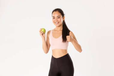 Spor, refah ve aktif yaşam tarzı konsepti. Zayıf ve sağlıklı bayan beden eğitimi hocası, spor eğitmeni vitamin yeme, baş parmağını kaldırma ve yeşil elma gösterme, gülümseme, beslenme önerisi.