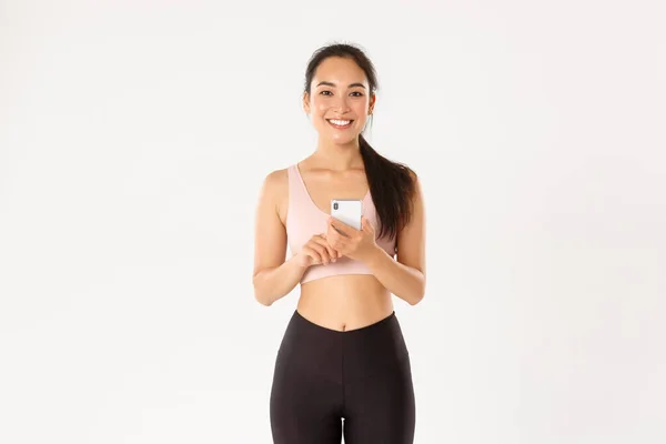 Esporte, tecnologia e conceito de estilo de vida ativo. Sorrindo satisfeito asiático fitness girl, sportswoman segurando smartphone, usando o aplicativo rastreador de execução, verificando heartrate durante o treino, fundo branco — Fotografia de Stock