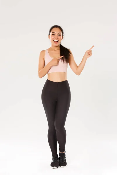 Longitud completa de atrevido atractivo femenino athelte, chica asiática de fitness en ropa deportiva señalando los dedos esquina superior derecha, demostrando promo, mostrando la venta de equipos de entrenamiento, descuento de entrenamiento de fitness —  Fotos de Stock