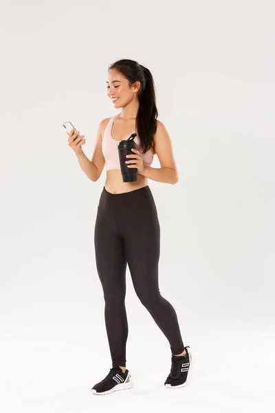 Comprimento total do ajuste sorridente e treinador de fitness feminino saudável, esportista asiático verificando lembrete de rastreador de água no smartphone, usando o aplicativo durante o treino no ginásio, fundo branco — Fotografia de Stock