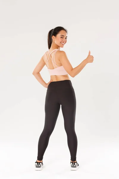 Longitud completa de motivado y satisfecho sonriente asiático entrenador femenino, chica athelte en ropa deportiva vuelta atrás para mostrar los pulgares hacia arriba en la cámara, listo para el entrenamiento de fitness productivo, fondo blanco — Foto de Stock