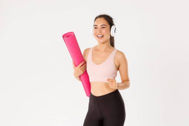 Spor, refah ve aktif yaşam tarzı konsepti. Neşeli gülümseyen Asyalı fitness kızı kulaklıklı ve spor giyimli lastik paspasla spor salonuna gidiyor, yoga dersleri için acele et heyecanlı, beyaz arka plan